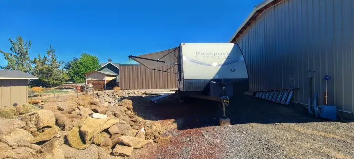 A 'Passport' travel trailer, great for an RV rental, is parked with its awning extended by a metal building. Dry sod is in the sunny foreground.. Keystone Passport 2016