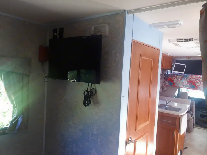 RV rental interior: a cozy living space featuring wallpapered walls, mounted TV, kitchen, and a bed over the cab.. Other Other 2009