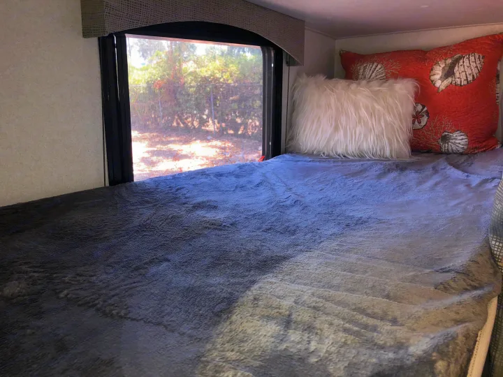 Inside an RV rental: a cozy sleeping area featuring a bed with a grey blanket, white furry pillow, and seashell pillow by a window.. Forest River Evo 2019