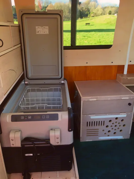 Inside an RV rental: An open Dometic portable fridge, showing its wire basket, sits next to a custom wooden power unit. Green field visible outside.. Toyota Hiace 2006