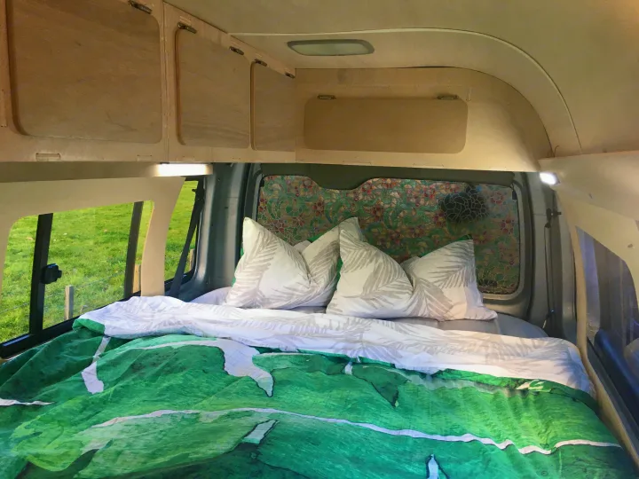 Inside a comfortable RV rental campervan, featuring a cozy bed with green bedding, overhead storage, and a field view.. Toyota Hiace 2006