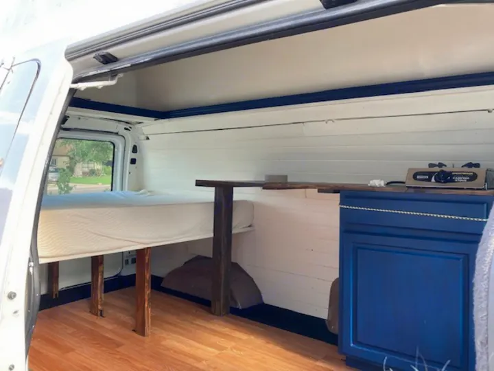 RV rental van conversion interior featuring a cozy bed, wood floor, and blue kitchen cabinet with camping stove.. Ford E350 Box Van 1997
