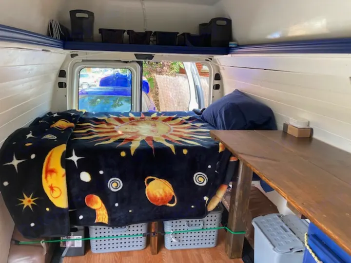 Cozy camper van interior, ideal for an RV rental. Features a large bed with a unique space-themed blanket, storage, and a handy table.. Ford E350 Box Van 1997