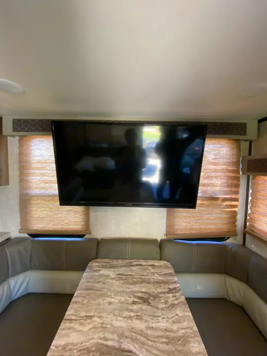 An RV rental's interior: wall-mounted TV, adjacent dining booth with patterned table, and window blinds.. Forest River FR3 2021