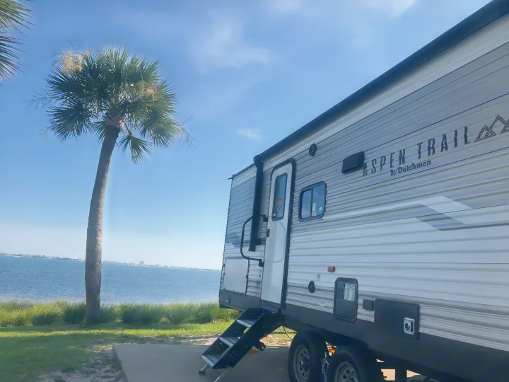 Enjoy your RV rental! An Aspen Trail travel trailer overlooks a calm bay with a palm tree, perfect for an outdoor escape.. Dutchmen Aspen Trail 2021