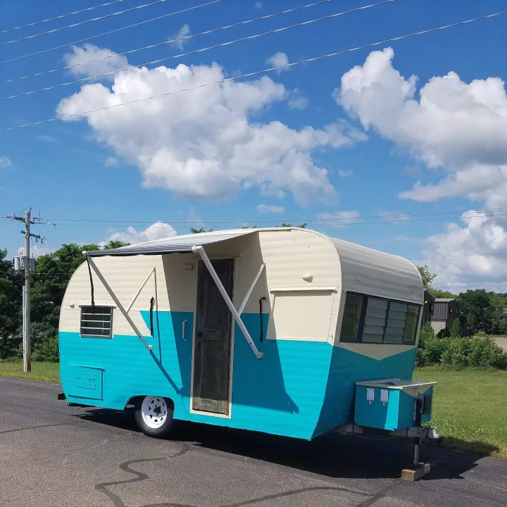A retro turquoise and cream RV rental, featuring an extended awning, sits on asphalt under a vibrant blue sky with clouds.. Nomad Rear fold 1962