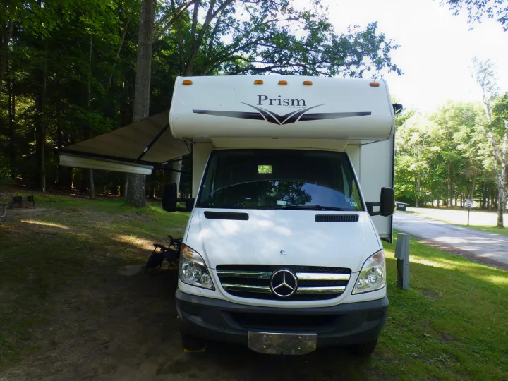 White RV rental parked at a wooded campsite with its awning extended, surrounded by trees and a road.. Coachmen Prism 2150LE 2013