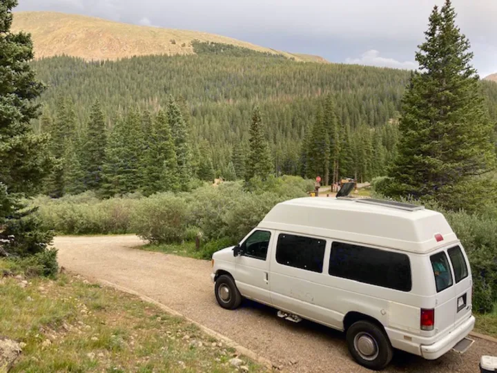 An RV rental van, white with a high top and solar panels, parked on a gravel path in a dense mountain forest.. Ford E350 Box Van 1997