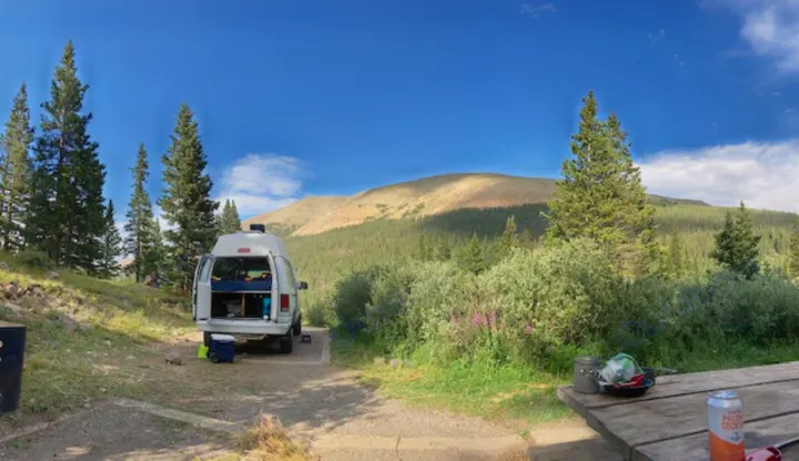 RV rental adventure! Camper van parked amidst tall trees, with open back doors, facing majestic mountains & forest. Ideal for camping.. Ford E350 Box Van 1997