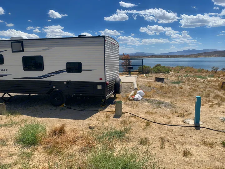 RV rental parked at a scenic lakeside campsite with mountain views under a blue sky. Power hookup visible.. Keystone Springdale 2021