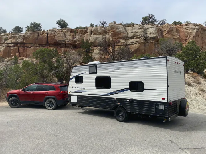 Red SUV towing a white Springdale travel trailer RV rental, parked near rocky cliffs. Ready for a scenic road trip!. Keystone Springdale 2021