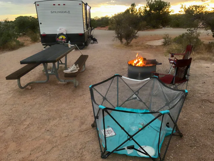 Relaxing at an RV rental campsite: fire pit, picnic table, chairs, and a playpen under a golden sky.. Keystone Springdale 2021