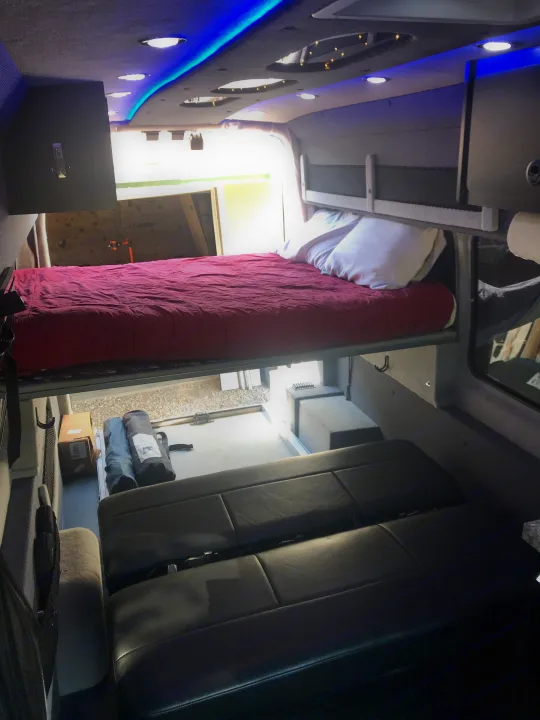 Interior view of an rv rental camper van, featuring a bed with red bedding, black passenger seats, and rear storage. Blue LED lighting.. Ford Transit Custom 2015