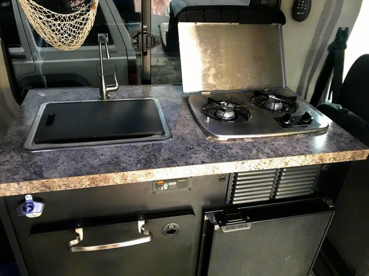 RV rental kitchen: a countertop with a sink (covered) and two-burner stove. Below, a Nova Kool fridge, pull-out table, and bottle opener.. Ford Transit Custom 2015
