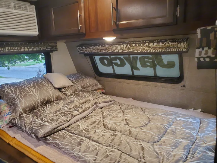 Cozy RV rental bedroom with patterned bedding, an AC unit, and overhead cabinets, offering comfortable travel accommodations.. Jayco Jay Flight 2019