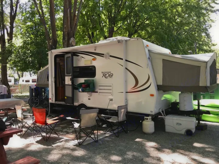 A white Rockwood Roo RV rental is parked at a gravel campsite under trees, with camping chairs and a grill outside.. Rockwood Roo 2011
