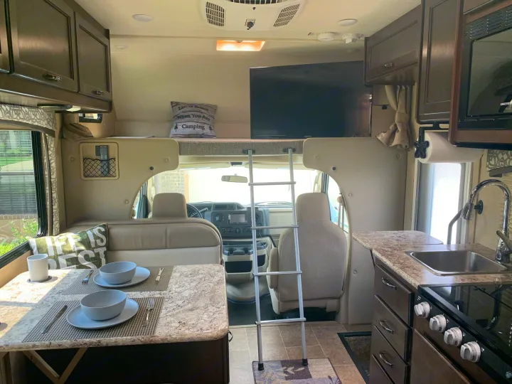 Well-equipped RV rental interior showcasing a dining area, kitchen, and overhead bunk with TV and ladder for travel.. Thor Chateau 2019