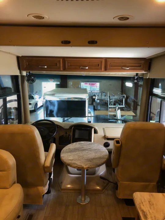 RV rental cockpit view: Spacious driver/passenger seats, small table, and dashboard looking out to a workshop.. Thor Motor Coach Challenger 2018