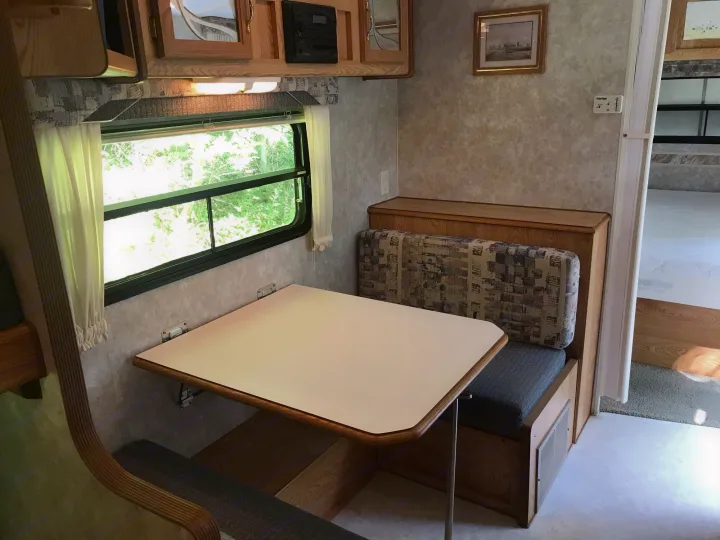 Cozy RV rental interior: A dinette with a table, booth seating, overhead cabinets, and a window overlooking lush greenery.. Sunline Solaris/Delivery&Set-up 2002