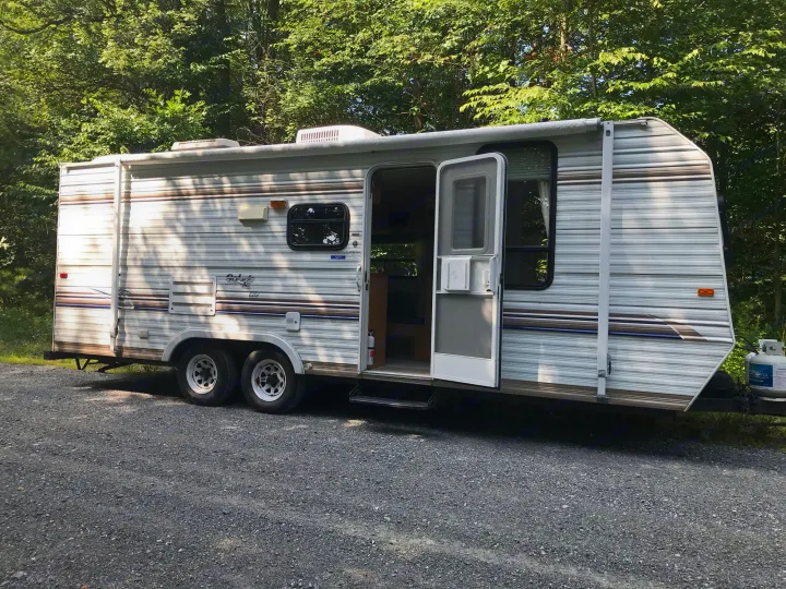 White travel trailer RV rental parked on gravel with its door open, surrounded by green trees. Ideal for outdoor adventures.. Sunline Solaris/Delivery&Set-up 2002