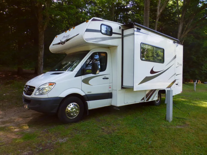 White Class C RV rental with slide-out extended, parked on grass at a shady campsite. Ideal for outdoor adventures.. Coachmen Prism 2150LE 2013