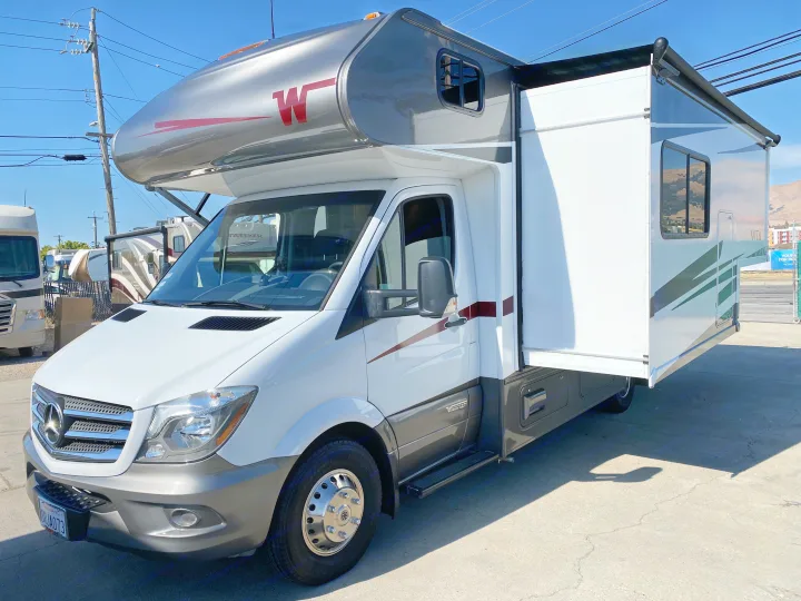 A white and grey Mercedes-Benz RV rental, a Class C motorhome, with its side slide-out extended on a lot.. Winnebago Vita 2020