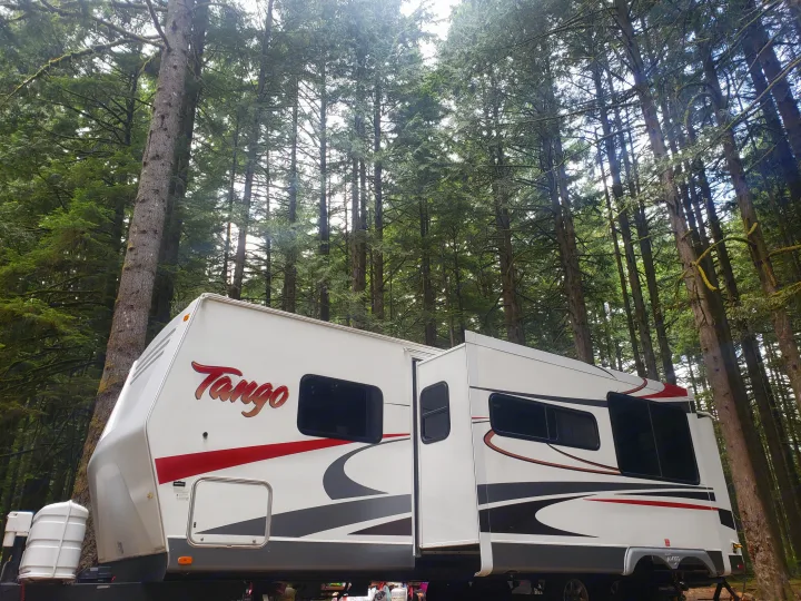 A white Tango RV rental, with red and gray accents, is nestled among tall, sunlit evergreen trees in a serene forest setting.. Pacific Coachworks Tango 2010