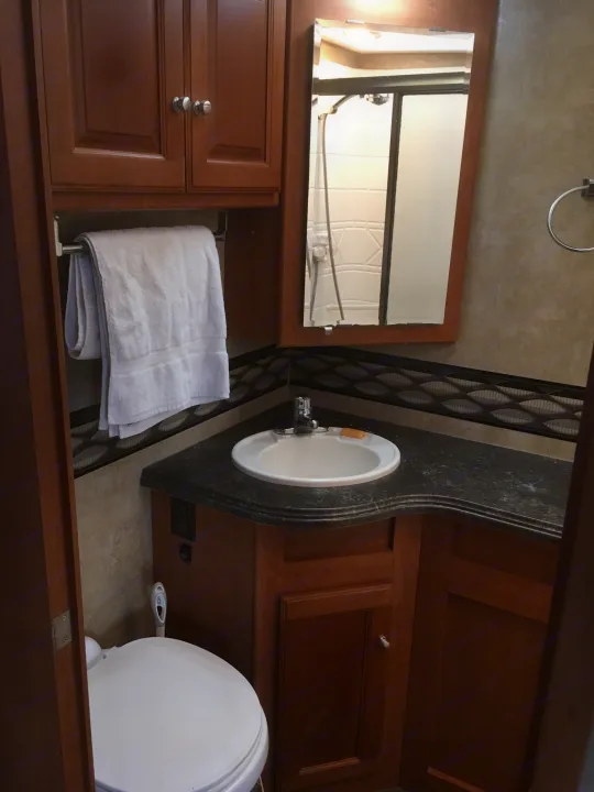 Clean RV rental bathroom with sink, mirror reflecting the shower, toilet, wood cabinets, and towel.. Winnebago Itasca Sunova 2014