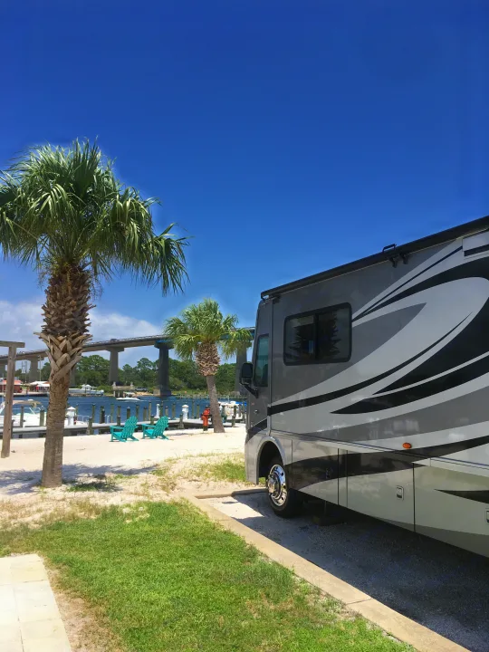 A spacious RV rental is parked by a sunny beach with palm trees, a pier, and a bridge in the distance, offering a scenic waterfront view.. Winnebago Itasca Sunova 2014