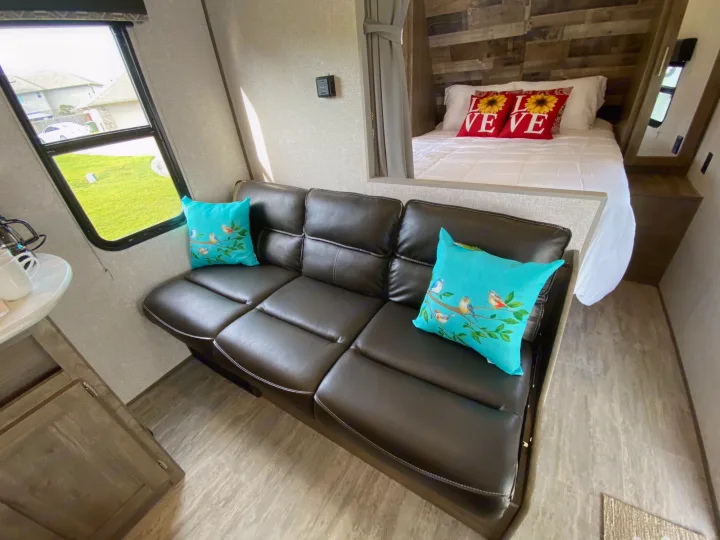Cozy RV rental interior features a dark leather sofa, colorful pillows, and a bedroom with a wood-paneled wall.. Dutchmen Aspen Trail 2021