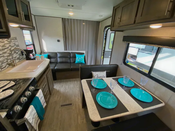 Inside an rv rental: modern kitchen with sink & stove, sofa lounge, and dining booth set with dishes. Cozy travel home.. Dutchmen Aspen Trail 2021