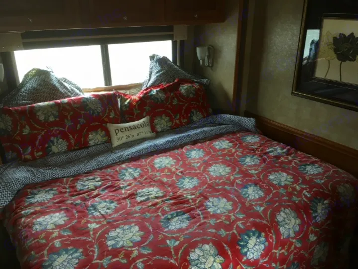 Cozy bedroom in an RV rental, featuring a red floral comforter and a decorative "Pensacola" pillow.. Winnebago Itasca Sunova 2014