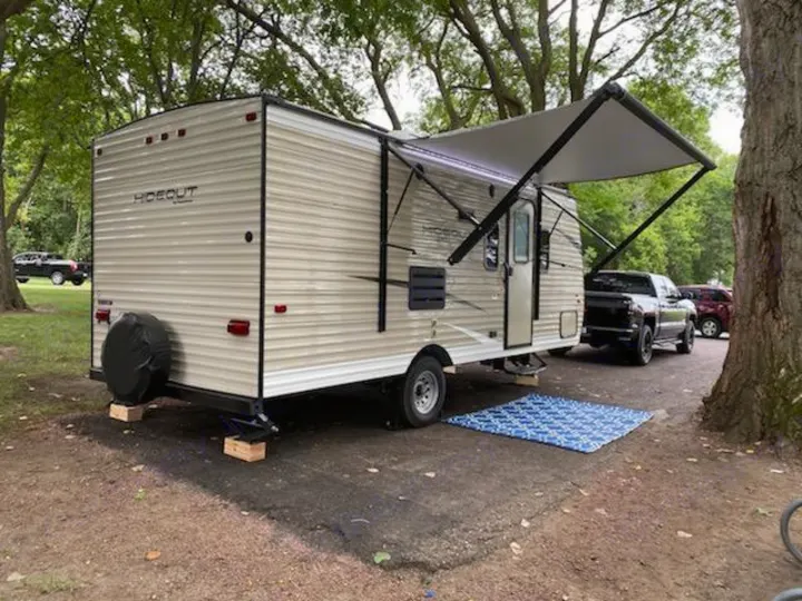 This RV rental, a beige travel trailer, is parked and set up with an awning extended at a shaded campsite.. Keystone Hideout 2019