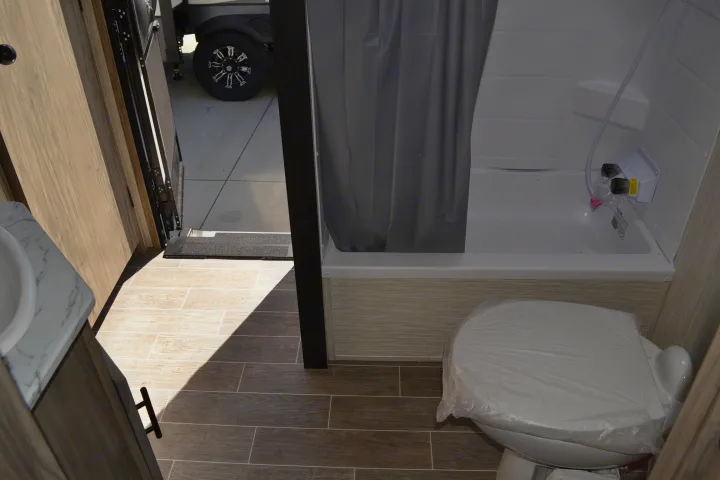 Inside an rv rental: Compact bathroom with white toilet (covered), shower/tub, sink, and wood-look floor. View to exit.. Forest River Other 2020