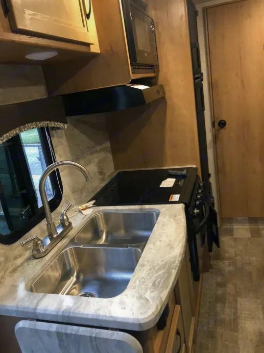 Compact RV rental kitchen with dual stainless steel sink, black stovetop/oven, microwave, and wood cabinets.. Winnebago Minnie Winnie 2021