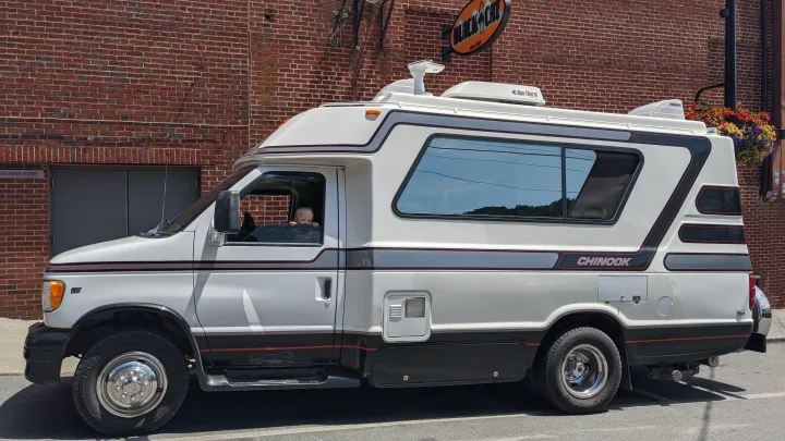 A white Chinook rv rental, a vintage camper van, is parked by a brick wall. A child peeks out the driver's window.. Chinook Concourse 2001
