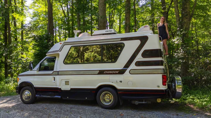 Woman on ladder by a white Chinook rv rental in a sunny forest. Ideal for nature adventures and camping.. Chinook Concourse 2001
