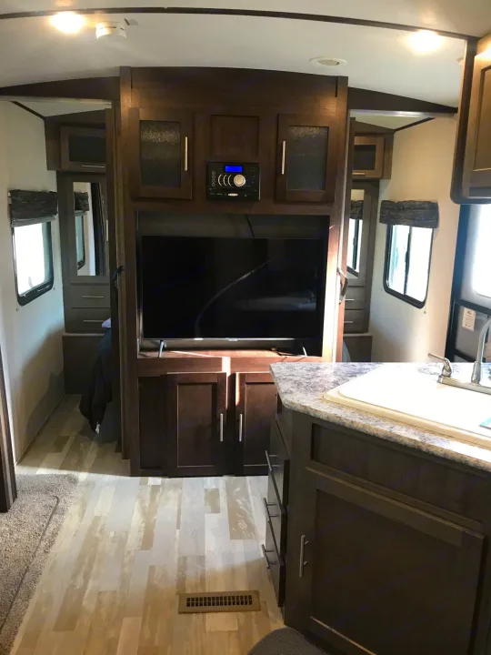 Inside a modern RV rental: a cozy living space with a TV, dark wood cabinetry, and a compact kitchen area.. Forest River Surveyor bunkhouse 2018