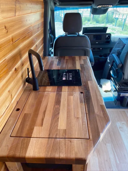 Modern RV rental interior with wood walls, sleek kitchen counter, sink, and induction cooktop. Perfect for camper van adventures!. Mercedes-benz 4x4 Sprinter mwb 2019