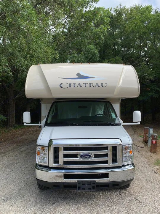 Front view of a white Ford Chateau RV rental, parked at a gravel campsite with utility hookups among trees.. Thor Chateau 2019