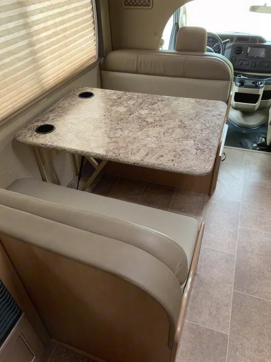 Inside an RV rental: dinette with table, built-in cup holders, comfy benches, and pleated window blinds.. Thor Chateau 2019