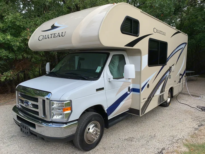 This white and tan Ford Chateau RV rental is parked on gravel with power and water connected, surrounded by trees.. Thor Chateau 2019