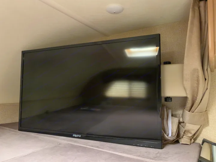 An Axxera flat-screen TV, part of an RV rental, rests on a cushioned surface, reflecting a light and blinds.. Thor Chateau 2019