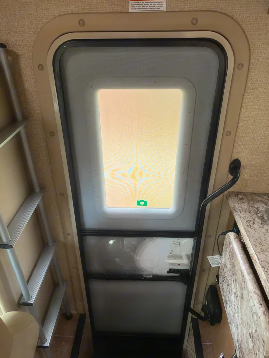 Inside an RV rental, a screen door with a bright outdoor view, a ladder for bunks, and a functional kitchenette are visible.. Thor Chateau 2019