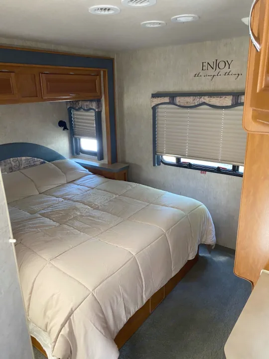 Cozy RV rental bedroom with a comfortable bed, overhead storage, and window. "Enjoy the simple things" decal adds charm.. Fleetwood Jamboree 2005