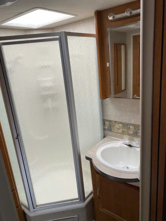 RV rental bathroom features a compact shower with frosted doors, a vanity sink, mirror, and ceiling skylight.. Fleetwood Jamboree 2005