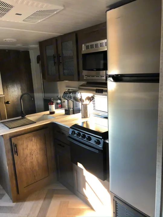 Modern RV rental kitchen with stainless steel fridge, microwave, stove, sink, and warm sunlight.. Forest River Wildwood X-Lite 2020