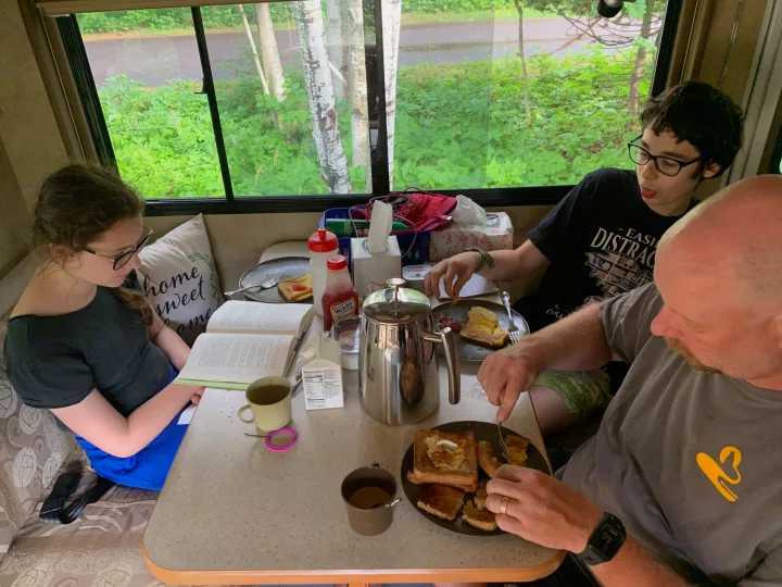Family enjoys breakfast with French toast and coffee inside their RV rental, while one reads a book.. Winnebago Access 2008