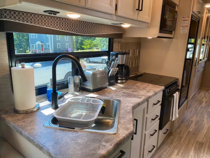 Modern rv rental kitchen featuring a large sink, black faucet, toaster, coffee maker, and full stove/oven. Bright interior.. Thor A.C.E 2020