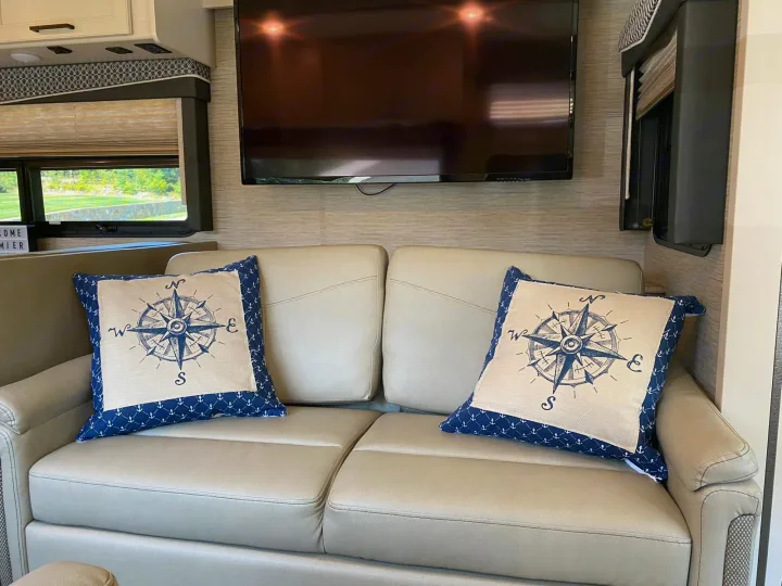 Inside an RV rental: a cozy sofa with compass-themed pillows and a TV. Ready for relaxing journeys.. Thor A.C.E 2020